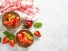 Fotografia em tons claros e vista de ciam de dois copos decorativos de tamanho normal com a sobremesa de taça moranguda, finalizada com chocolate e decoração de morangos em pedaços e folhas de hortelã.