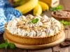 Em cima de uma mesa, banoffe com massa crocante, decorado com chantilly cremoso, bananas cortadas em rodelas e chocolate ralado.