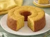 Bolo de milho dourado e fofinho, inteiro com fatia cortada, servido em prato branco sobre mesa rústica.