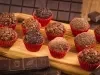 Fotografia em tons de vermelho em uma bancada de madeira com vários brigadeiros colocados em forminhas vermelhas em cima de uma tábua de madeira clara. Ao fundo, uma barra de chocolate meio amargo e com alguns pedaços de chocolate espalhados.
