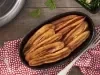 Fotografia em tons de vermelho em uma bancada de madeira, uma toalha vermelha com flores, um recipiente oval preto com várias bananas fritas dentro dele.