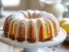 Fotografia de um bolo bundt dourado em um suporte branco, coberto com glacê branco e polvilhado com açúcar de confeiteiro. O fundo apresenta um bowl com limões amarelos.