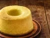 Fotografia de um bolo de milho recém-assado, com formato de bundt, dourado e macio, colocado em uma tábua de madeira. O fundo rústico.