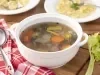Fotografia em tons de vermelho em uma bancada de madeira, um pano xadrez vermelho, uma panela branca funda com alças laterais com o caldo de legumes dentro dela. Ao lado, uma concha e dois pratos brancos fundo com sopa de capelleti com o caldo de legumes.