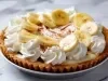 Fotografia em tons claros de uma banoffee inteira e completa dentro de um recipiente meio fundo sobre uma mesa de mármore.