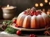 Fotografia de um bolo de Natal em um prato redondo, polvilhado com açúcar de confeiteiro e adornado com frutas vermelhas e pinhas. Velas acesas ao fundo proporcionam um ambiente aconchegante.