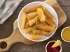 Imagem vista de cima de um prato branco com mandioca frita sobre uma tábua de madeira. Ao lado, um potinho de ketchup e um de mostarda.