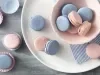 Imagem vista de cima de macarons rosa e azul em um pote rosa claro e ao lado, macarons em forminhas de papel.