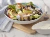 Fotografia em tons de branco e azul de uma bancada branca e uma tábua de madeira, sobre ela um recipiente redondo com a salada e um garfo. Ao lado uma molheira branca com o molho e um paninho branco e azul.