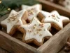 Em cima de uma mesa, biscoitos de estrelas, decorados com glacê branco e confeitos dourados de bolinha.