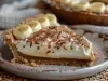 Em cima de uma mesa, fatia de banoffee com massa de biscoitos crocante, doce de leite, decorado com chantilly cremoso, banana em rodelas e cacau em pó.