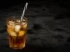 Fotografia em tons escuros de um copo de vidro com uma bebida de guaraná com cubos de gelo e um canudo preto e branco dentro do copo, o qual está sobre uma mesa preta.