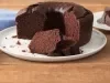 Fotografia em tons de marrom e branco de uma bancada de madeira clara com um paninho azul claro e um prato branco redondo com o bolo de chocolate. Aos lados dois pratos brancos pequenos com fatias de bolo de chocolate e um garfo.