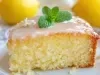 Fotografia de uma fatia de bolo de limão em um prato decorativo. O bolo apresenta uma textura fofa, coberto com glacê brilhante e decorado com hortelã, cercado por limões inteiros e flores brancas.