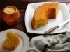 Fotografia de um bolo de fubá parcialmente fatiado em um prato branco, com uma crosta dourada e interior amarelo. Um recipiente de madeira com fubá está ao lado.