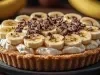 Em cima de uma mesa, banoffee com massa de biscoitos crocante, chantilly cremoso, bananas em rodelas, polvilhado com rapas de chocolate.