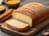 Fotografia de um bolo recém-assado em uma tábua de madeira, com uma crosta dourada e polvilhada com açúcar. Ao fundo, especiarias e ingredientes em pequenas tigelas.
