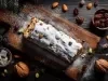 Fotografia de um bolo em uma tábua rústica, coberto com açúcar de confeiteiro. Acompanhado de nozes, amêndoas e um bowl de passas, a composição destaca tons quentes e texturas naturais