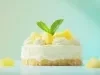 Fotografia em tons claros de uma mini torta de abacaxi decorada com duas folhas de hortelã, o fundo tem tom verde água e tem alguns cubinhos de abacaxi em volta da torta.