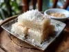 Fotografia de um prato de sobremesa tradicional, com pedaços retangulares de bolo de tapioca cobertos com coco ralado em um prato decorativo. O fundo desfocado sugere um ambiente natural.