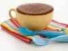 Foto de uma caneca bege com bolo dentro em cima de um pano colorido e com 2 colheres azuis do lado