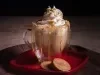 Foto da receita de cappuccino caramelo salgado servida em dois copos de vidro sobre uma mesa de madeira com uma mão polvilhando caramelo salgado