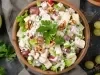 Fotografia de uma salada vibrante em uma tigela de madeira. A mistura inclui cubos de batatas cozidas, maçã-verde, uvas roxas e nozes, coberta com um molho cremoso e decorada com salsa, destacando frescor e texturas harmoniosas.