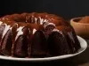 A imagem apresenta um bolo de chocolate Bundt, disposto em um prato cerâmico branco. A cobertura de chocolate brilhante escorre suavemente pelas laterais do bolo. A textura do bolo parece densa e úmida.