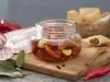 Fotografia em tons de vermelho em uma bancada de madeira clara, um pano branco com listras vermelhas, uma tábua de madeira com um pote hermético de vidro em cima e com as pimentas em conserva dentro dele. Ao fundo, uma cesta com pasteis e pimentas.