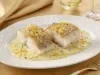 Filés de bacalhau servidos com molho cremoso de ervas e raspas de limão, apresentados em prato branco elegante.