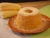 Imagem mostra bolo de milho redondo com furo no meio em um prato vermelho e ao lado de de espigas de milho