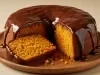 Bolo de cenoura em forma de anel, com a mesma cobertura generosa de chocolate escorrendo. Uma fatia já foi retirada e está deitada ao lado, mostrando a textura úmida e fofa da massa.