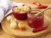 Fotografia em tons de roxo em uma bancada de madeira clara, um pano branco com lilás xadrez, uma tábua de madeira clara com um potinho de vidro e a geleia de pimenta dentro. Ao lado, um potinho vermelho com os dadinhos de tapioca.