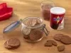 Fotografia de um potinho de vidro com tampa sobre uma mesa de madeira clara, ao lado da lata de NESCAU Extra Cacau 30% e um copo de vidro com leite e nescau. Dentro e em volta do pote tem os biscoitinhos amanteigados de nescau.