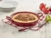 Imagem de uma caçarola vermelha com polenta e carne moída sobre um paninho xadrez vermelho e branco. Compondo a imagem, uma colher, manjericão, tomates e pratos brancos.