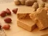 Fotografia em tons de caramelo com uma paçoca de formato retangular ao centro. Em volta da paçoca existe alguns amendoins