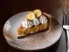 Em cima de uma mesa, fatia de banoffee com massa de biscoitos crocante, doce de leite, bananas em rodelas, decorado com chantilly cremoso, banana em rodelas e cacau em pó.