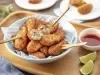 Fotografia em tons de bege em uma bancada de madeira clara, uma tábua de madeira, um pano azul, um prato redondo fundo com vários bolinhos de peixe, um potinho branco pequeno com molho de pimenta e pedaços de limão. Ao fundo, pratinhos brancos com bolinho