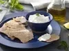 Imagem de um prato azul com um bowl com coalhada seca e pedaços de pão sírio ao lado. Ao fundo, um vidro de azeite e pedaços de limão.