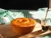 Um bolo de cenoura inteiro, com formato redondo e com furo no meio, está sobre uma tábua de madeira. Ao lado, um copo está sendo servido com café. O fundo tem um pano de prato verde e branco.