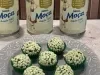Docinhos verdes com granulado branco, em forminhas verdes sobre prato cinza, com latas de Leite Moça ao fundo.