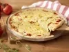 Fotografia em tons de vermelho em uma bancada de madeira clara, uma tábua de madeira, um pano branco com listras vermelhas, uma forma de pizza com a pizza de muçarela em cima dela e uma espátula de madeira tirando um pedaço. Ao lado, tomates para decorar.