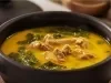 Receita de frango servido no caldo de tucupi com folhas de jambu. Uma foto bem aproximada que mostra uma panela de barro preta com tudo dentro.