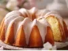 Fotografia de um bolo bundt dourado, apresentado em um prato decorativo. O bolo possui uma cobertura branca brilhante, levemente polvilhada com açúcar de confeiteiro, em um fundo suave com elementos florais.