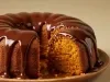 Um bolo de cenoura assado em forma de anel, com cobertura espessa de chocolate brilhante escorrendo pelas laterais. O bolo está parcialmente cortado, revelando sua textura macia e alaranjada no interior.