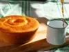 Um bolo dourado, assado em forma redonda com furo no meio, está sobre uma tábua de madeira. Ao lado, há uma caneca branca esmaltada sendo preenchida com café recém-passado. O fundo é composto por uma toalha xadrez verde e branca.