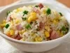 Em cima de uma mesa, tigela redonda com porção de arroz com presunto cortado, ervilha, cenoura picada e milho.