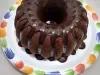 Foto em tons de marrom da receita de bolo de café servida com uma cobertura de chocolate por cima e sobre um prato branco com a borda colorida