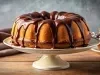 Fotografia de um bolo bundt dourado, coberto com um brilho de chocolate escuro, apresentado em um pedestal claro.