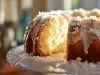 Fotografia de um bolo bundt dourado, exibido em um suporte de vidro decorativo. O bolo é coberto com uma cobertura branca que escorre pelas laterais, salpicado com flocos de coco.